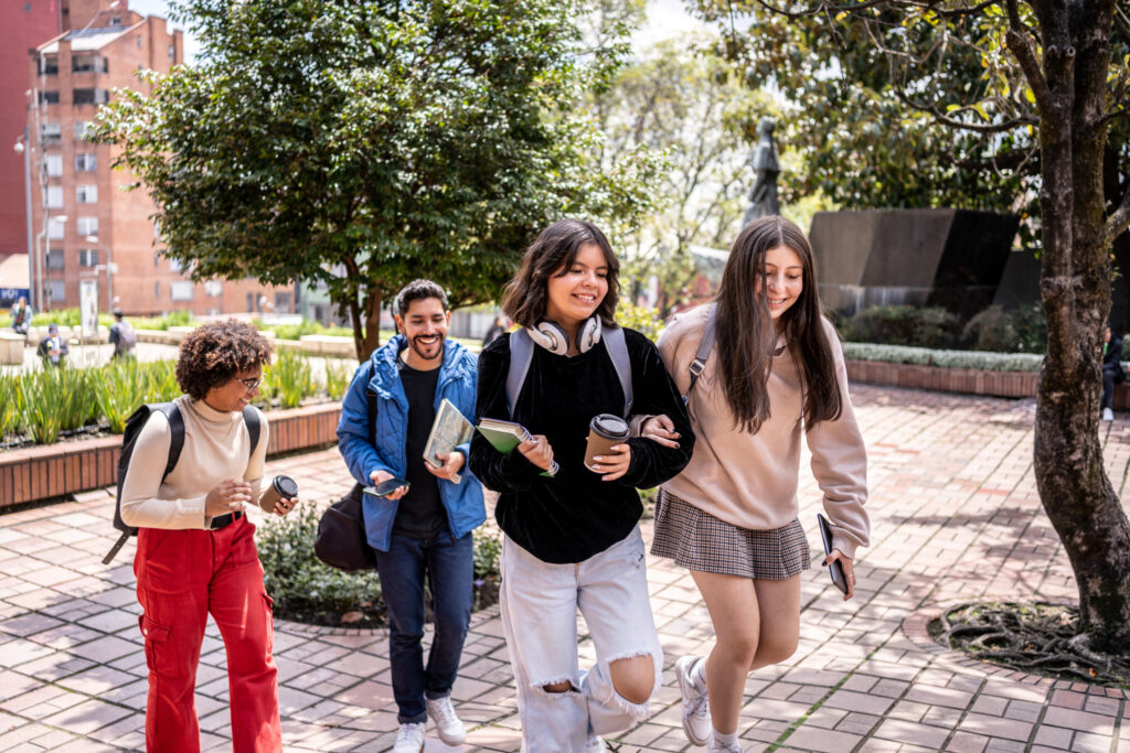 Four college students walking and laughing together on a brick path through campus, carrying books, drinks, and backpacks.