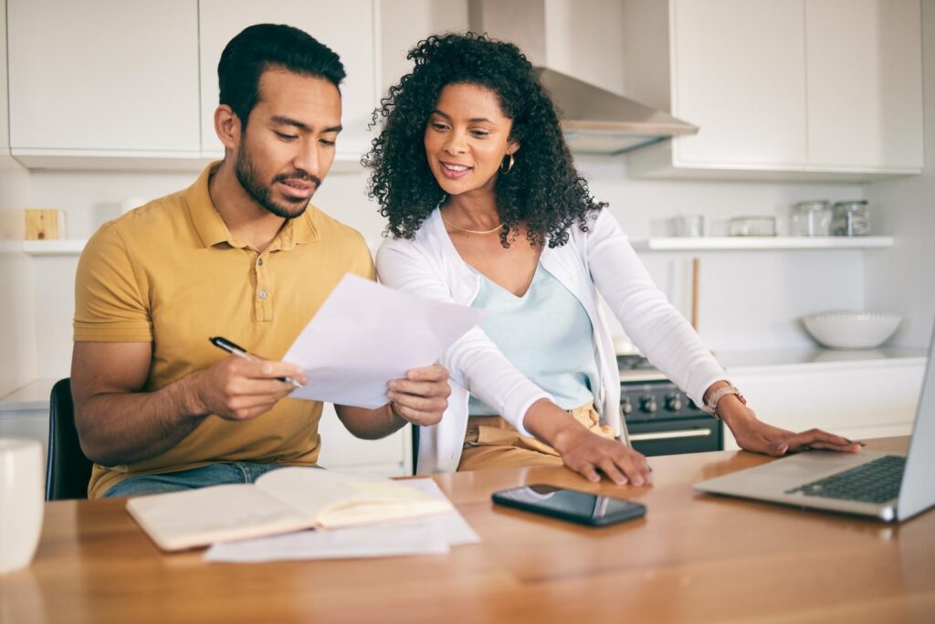 A couple reviewing finances together at a kitchen table, with a laptop, notebook, and phone in front of them.