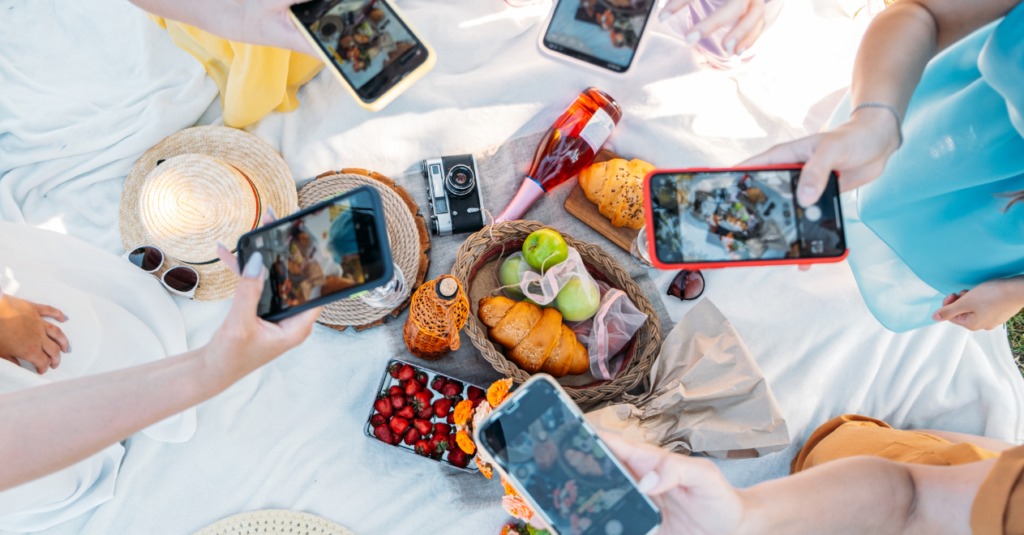 An overhead view of people photographing a picnic spread of fruit, pastries, and drinks with their smartphones.