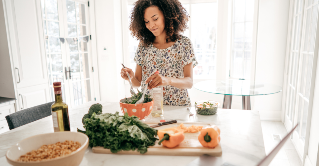 A woman with curly hair preparing salad in a bright kitchen, smiling with fresh vegetables on the counter