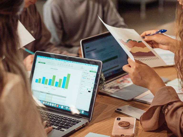 A group of people reviewing charts and data on laptops and printed reports during a collaborative meeting.