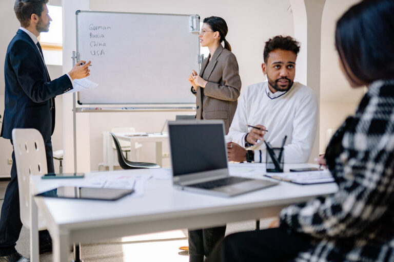 A diverse group of professionals collaborating in an office meeting, with two people presenting beside a whiteboard.